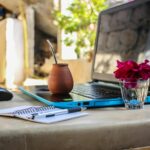 A serene outdoor workspace featuring a laptop, notebook, mug, and flowers on a desk.